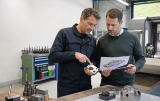Ein Techniker und ein Redakteur prüfen in einer österreichischen Werkstatt ein Metallbauteil anhand eines Entwurfs